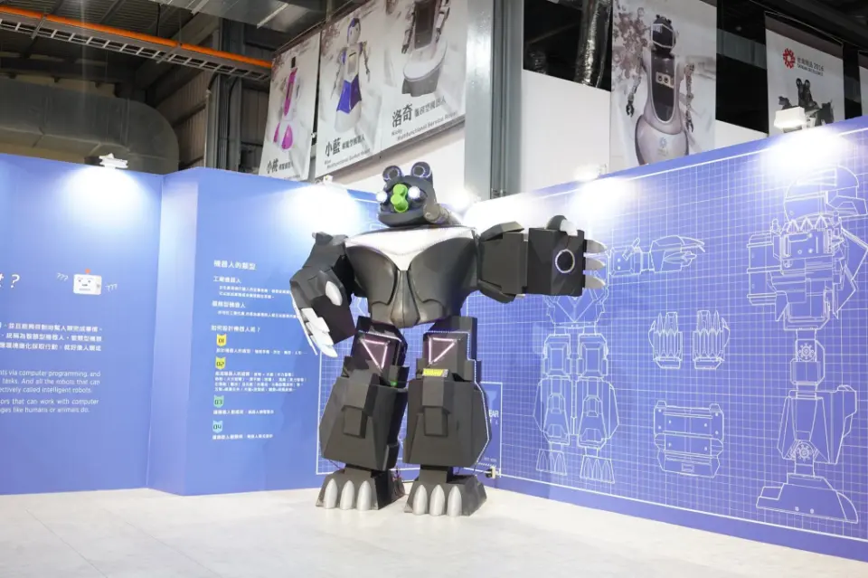 In Taoyuan Xiangyi Robot Dream Factory, a large black bear-shaped robot model is displayed with glowing purple accents, set against a blue wall featuring its design blueprints, showcasing future technology.