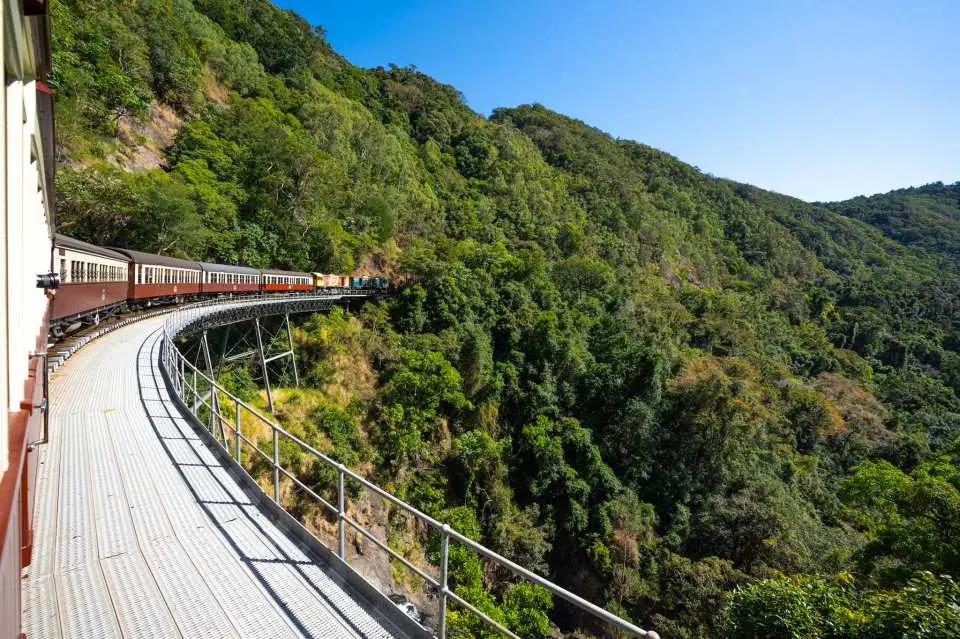 キュランダ観光列車の車窓から、列車が曲がりくねった線路に沿って豊かな熱帯雨林の斜面を走行する、美しい景色が見えます。