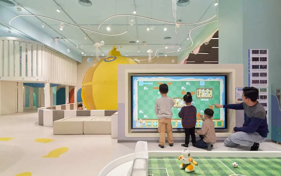 巧虎夢想樂園的數位互動體驗區，家長與小朋友在大型螢幕前玩體感遊戲，背景有黃色充氣設施，提供多元親子娛樂。