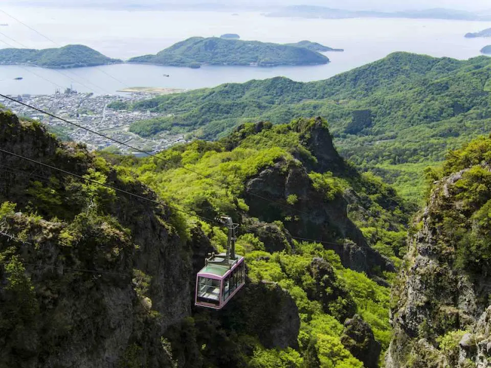 กระเช้าไฟฟ้าคันคาเคกำลังเคลื่อนที่เหนือภูเขาสีเขียวสดใสบนโชโดชิมะ คางาวะ ญี่ปุ่น พร้อมทิวทัศน์เมืองชายฝั่งและทะเลเซโตะที่กว้างใหญ่ภายใต้ท้องฟ้าสีครามสดใส