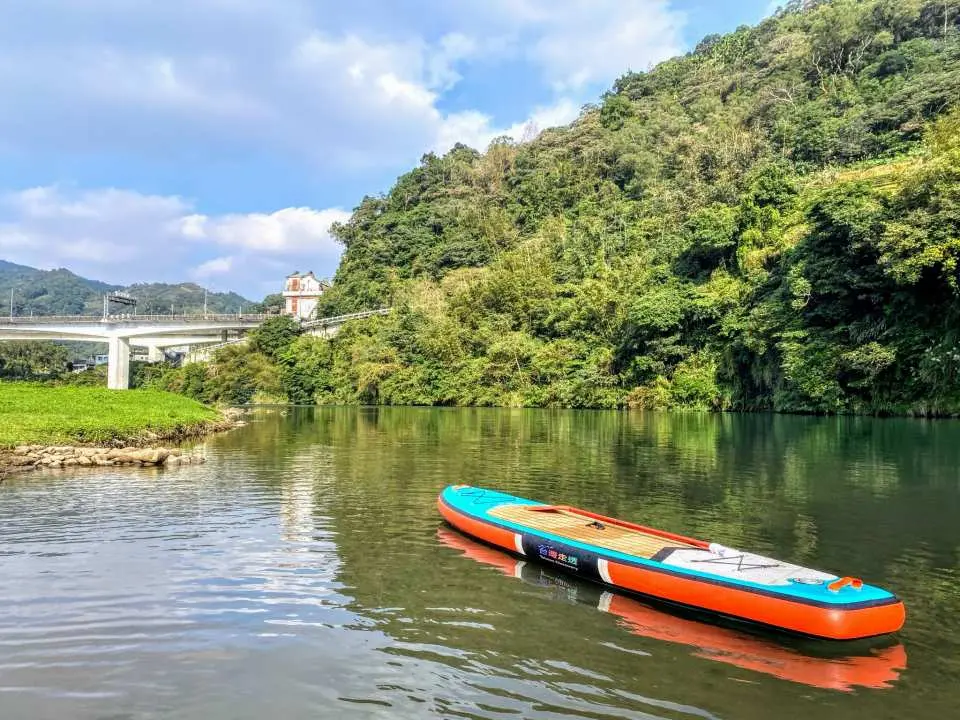 一艘色彩鮮豔的SUP板在清澈的坪林北勢溪上靜靜漂浮，周圍是翠綠的山丘和遠處的橋樑，彰顯寧靜的自然美景。