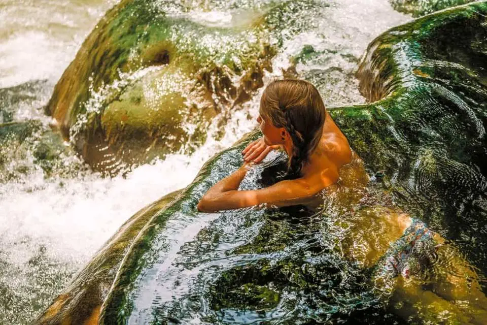 크라비 온천의 자연 풀에서 한 사람이 따뜻하고 맑은 치유 온천수에 몸을 담그고 있다. 주변은 녹색 이끼로 덮인 바위들로 둘러싸여 자연 속에서 휴식을 취하는 순간을 즐긴다.