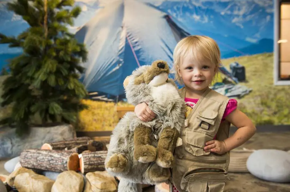 A joyful little girl in a safari vest, hugging a stuffed wolf, happily playing in a simulated outdoor camping scene at the Denver Children's Museum, offering interactive family fun.