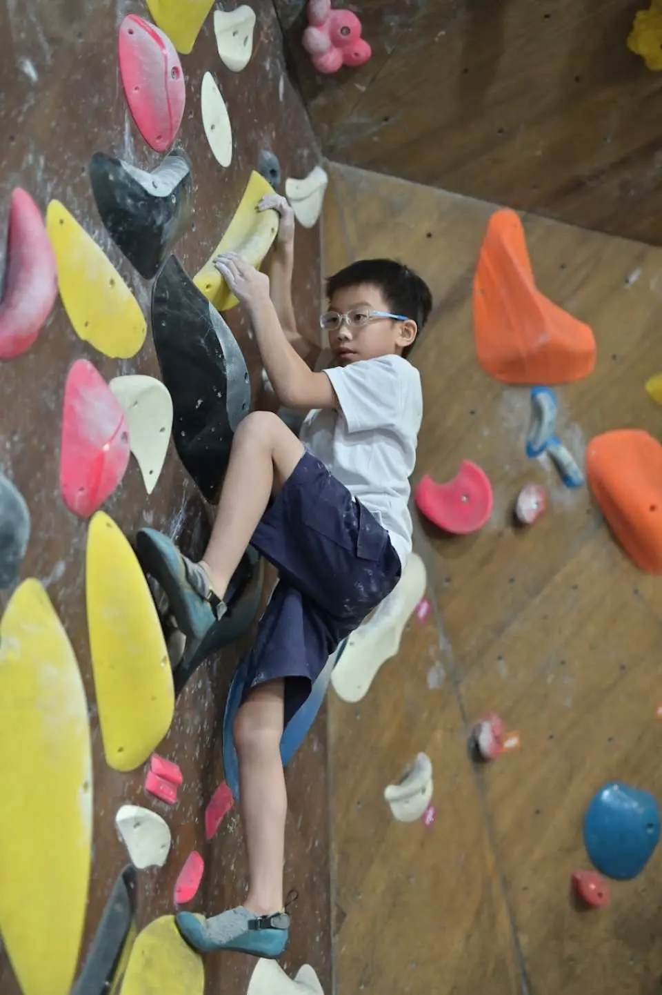 戴眼鏡的小男孩於JUST CLIMB室內抱石牆上積極攀爬，享受挑戰，體驗新手抱石課程的樂趣。