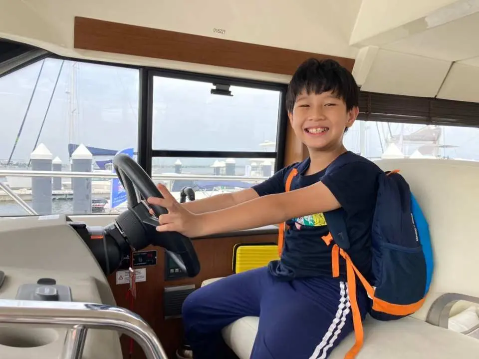 An excited young boy with a backpack in the cockpit of the Fangxing luxury yacht in Pingtung, Taiwan, happily experiencing the yacht driving fun, showcasing a joyful parent-child activity.