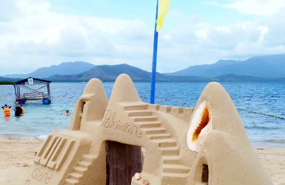 在巴拉望本田灣的露麗島，精美的沙雕上刻有「Luli Island Palawan」字樣和貝殼裝飾，背景是平靜的藍色海水和遠處的山巒。