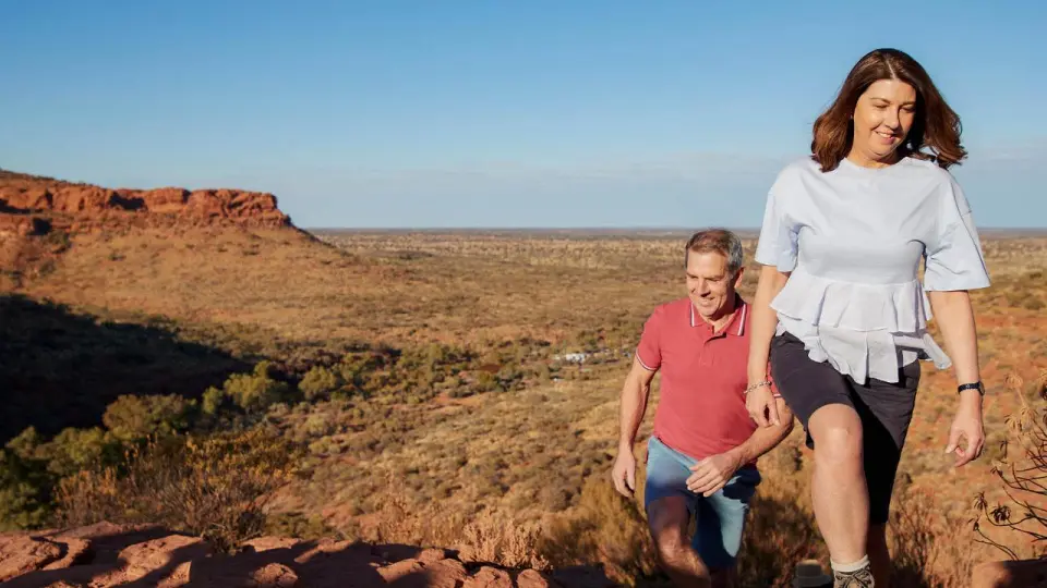 A couple of tourists energetically climbing uphill on the Kings Canyon Rim Walk, with the sun-drenched Australian outback scenery in the background, showcasing the vitality and joy of exploring nature.