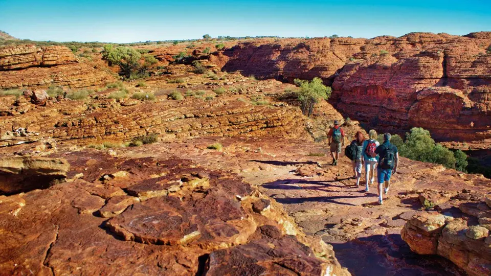 A group of adventurers hiking on the rugged red sandstone trail along Kings Canyon's rim, surrounded by unique canyon geology, enjoying the vastness and grandeur of the Australian outback.