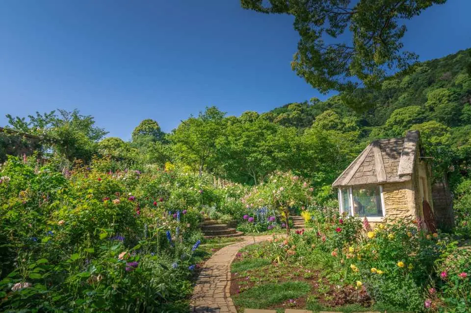 熱海ACAO FORESTの生命力あふれる赤尾ハーブ＆ローズガーデンの一景。曲がりくねった小道の両側には花々が咲き誇り、可愛らしい童話の小屋へと続き、庭園の幻想的な魅力を示しています。