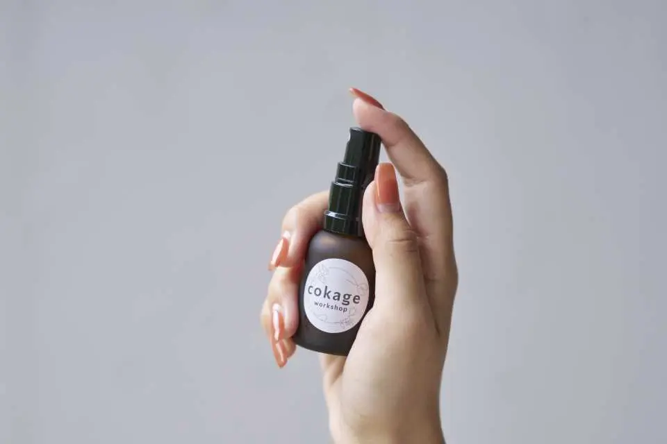 熱海ACAO FORESTのワークショップで作られた製品。手に持たれた「cokage」ラベルの付いた暗色ガラススプレーボトルは、手作りのハーブエッセンシャルオイルスプレーであり、天然素材の活用を示しています。