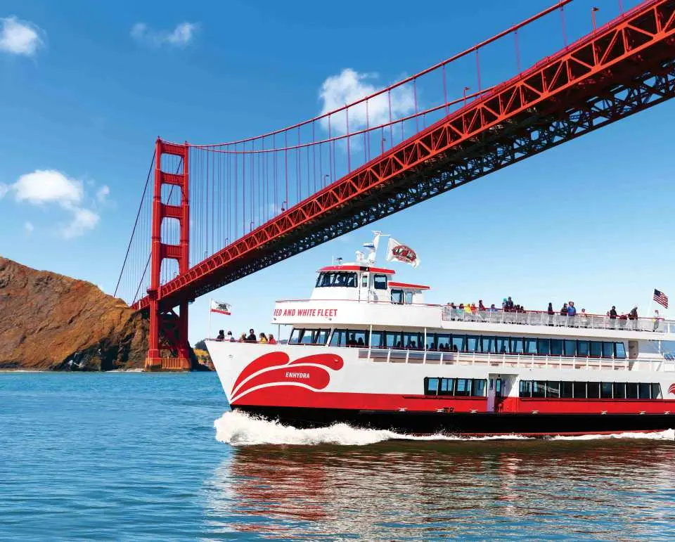 A Red and White Fleet sightseeing cruise boat sails beneath the Golden Gate Bridge, with clear blue skies and the vibrant bay, showcasing San Francisco Bay's iconic sightseeing route.