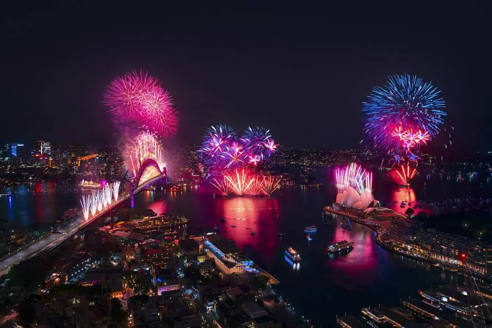 Aerial view of spectacular New Year's Eve fireworks exploding over Sydney Harbour Bridge, illuminating the Sydney skyline with vibrant colors, as cruise boats gather on the water.