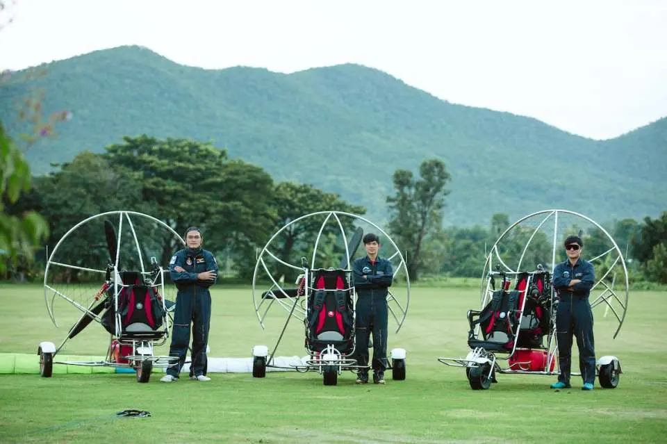 Three professional instructors in dark flight suits stand confidently in front of three powered paragliders on a vast green field in Chiang Mai, emphasizing expert guidance and safety.