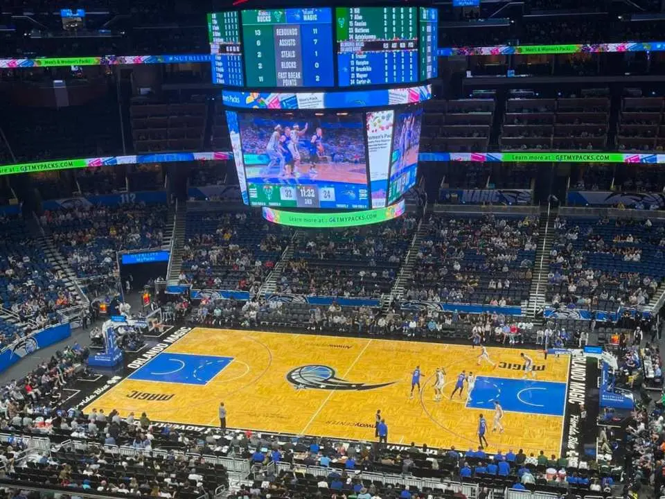 Elevated view of an Orlando Magic NBA home game, showcasing the court with players in action. The massive central jumbotron displays live game replays, enhancing the immersive basketball viewing experience for fans.