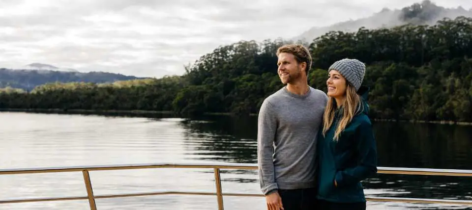A smiling couple on the deck of the Gordon River cruise, enjoying panoramic views of Tasmania's pristine wilderness and the tranquil river under a cloudy sky.