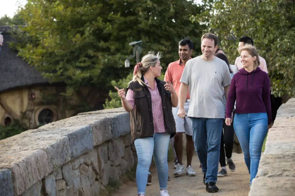A tour guide leading a group of visitors across a stone bridge within the Hobbiton movie set, exploring the charming Hobbit village with expert English commentary.