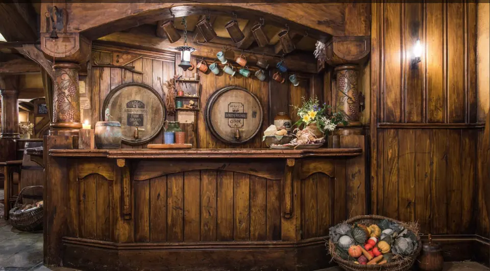 The rustic wooden interior of Hobbiton's Green Dragon Inn, featuring a cozy bar area with barrels, hanging mugs, and traditional decor, offering a charming place for visitors to relax.