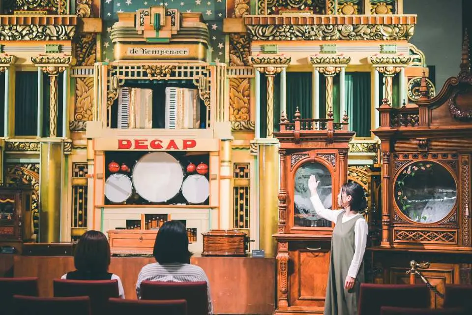 六甲オルゴールミュージアム内で、スタッフが大型の自動演奏楽器を訪問者に説明しています。神戸六甲山ならではの音楽展示と毎日開催されるコンサートを楽しめます。