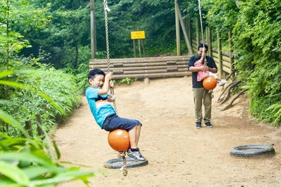 一名孩童在日本六甲GREENIA運動公園體驗獨特的橘色球形鞦韆，後方有家長帶著嬰兒，享受親子同樂時光。