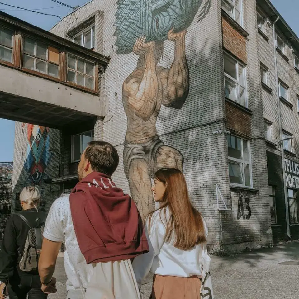 タリンのストリートアート壁画、巨大な鳥を抱える筋肉質の男性が描かれ、観光客がこのユニークな現代アート作品を鑑賞、都市文化とタリンカードの便利さを体験。