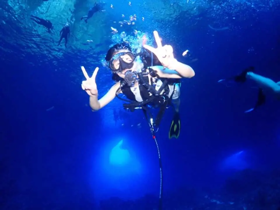 An excited diver in full scuba gear gives peace signs underwater in the brilliant blue depths of Saipan's Grotto, enjoying a safe introductory dive experience for all skill levels.