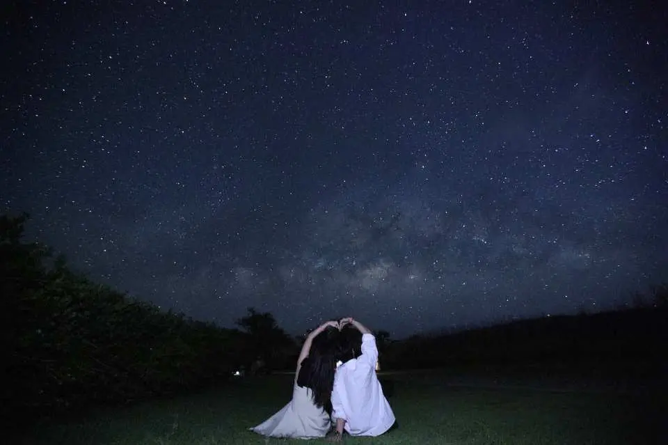 사이판 잔디밭에 앉아 서로를 안고 고요한 별이 빛나는 밤하늘을 올려다보는 커플이 스타라이트 투어에서 친밀하고 로맨틱한 경험을 나누는 모습.