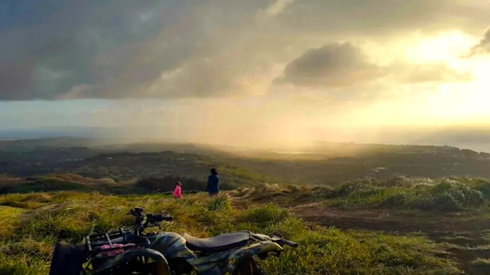サイパンのタポチョ山からの穏やかな夕日景色、ATVが草深い丘に停車し、霞がかった海岸線と海を見下ろし、島の自然の美しさを際立たせています。