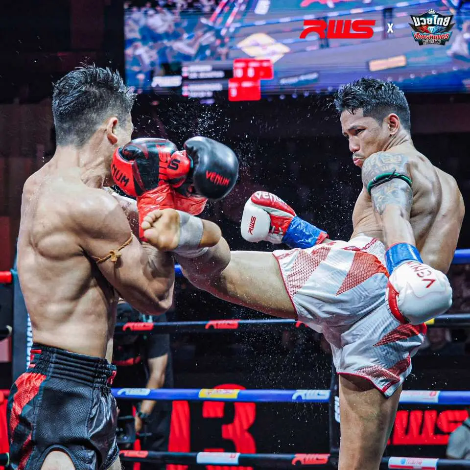 Hai võ sĩ Muay Thái ở Bangkok đang trao đổi những cú đấm và cú đá mạnh mẽ trên võ đài, thể hiện những pha giao tranh quyết liệt với găng tay màu đỏ và xanh lam rõ ràng trước mắt khán giả.