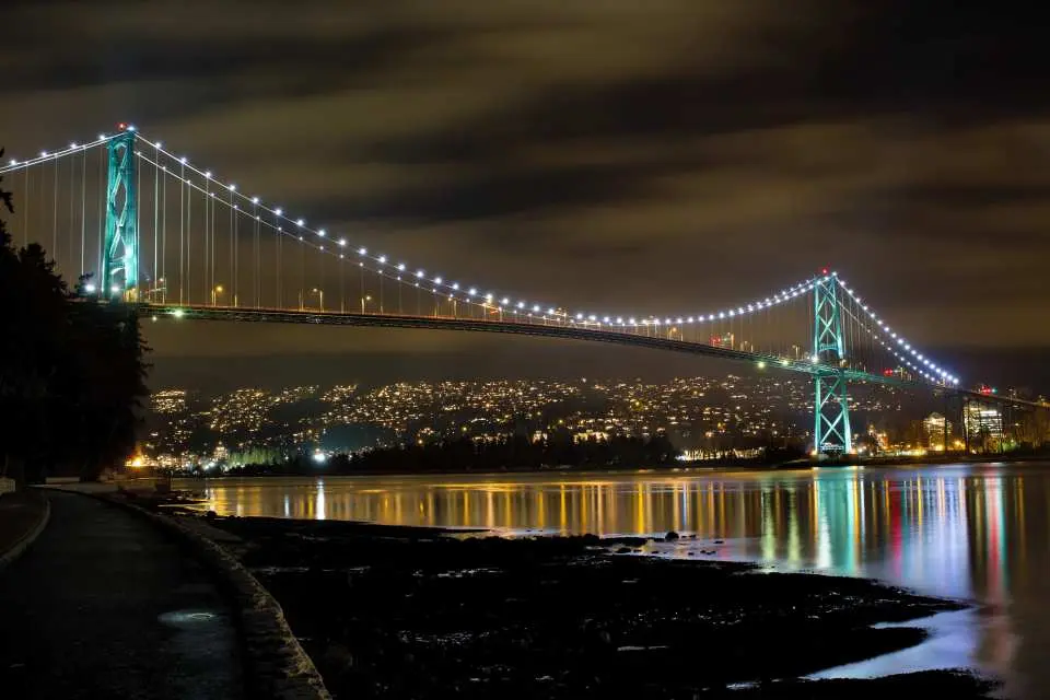 Vancouver's iconic Lions Gate Bridge at night, brilliantly illuminated, reflecting on the water, creating a captivating city skyline view.