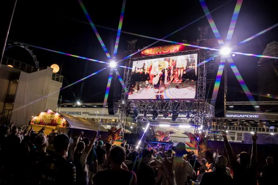 Vibrant outdoor live entertainment at the 2026 F1 Singapore Grand Prix, with attendees enjoying performances on a large screen and dynamic light beams illuminating the night sky.
