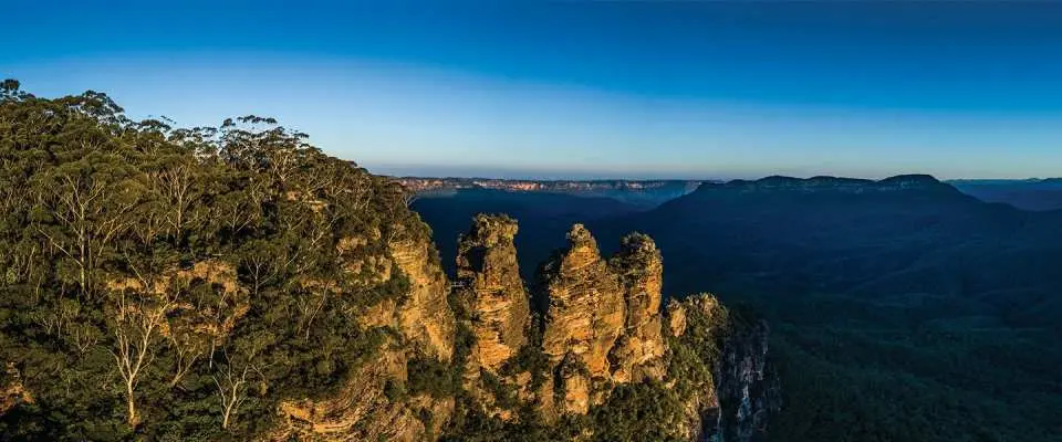 Toàn cảnh mỏm đá Ba Chị Em biểu tượng tại Dãy núi Xanh, trong ánh sáng vàng bình minh/hoàng hôn, nhìn ra thung lũng và rừng tươi tốt.