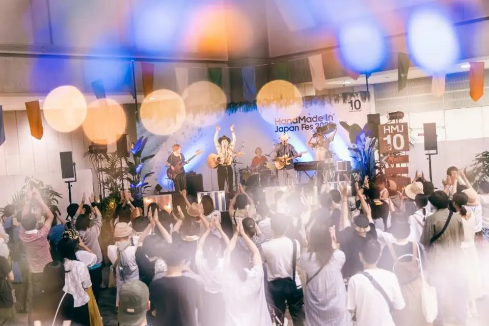 A band delivers an energetic live performance on stage in the music and entertainment area of HandMade In Japan Fes, with the audience waving their hands, fully immersed in the joyful atmosphere of Tokyo's largest creator festival.