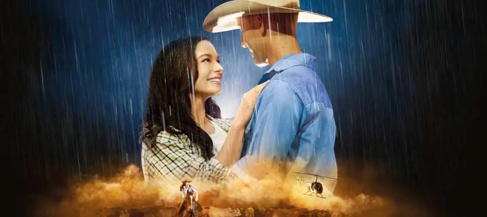 Ảnh nghệ thuật chính đầy kịch tính cho chương trình Australian Outback Spectacular, mô tả một cặp đôi ôm nhau dưới mưa cùng một cao bồi cưỡi ngựa và trực thăng phía dưới, nhấn mạnh câu chuyện giàu cảm xúc.