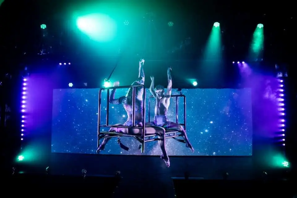 Two acrobatic performers execute an aerial act on stage at Dracula's 'Lucid' show in Gold Coast, against a starry background and dazzling lights, creating a stunning visual spectacle.