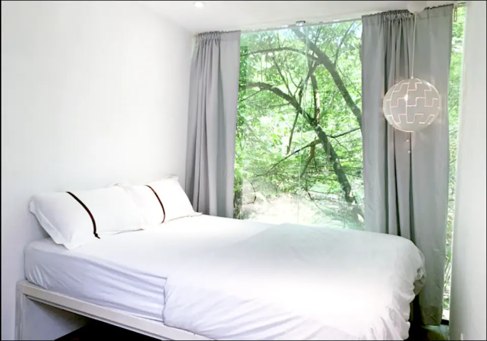 Modern and spacious room interior at Time Capsule Retreat, Sungai Lembing, featuring a comfortable white bed and a large window offering immersive views of the surrounding lush forest.