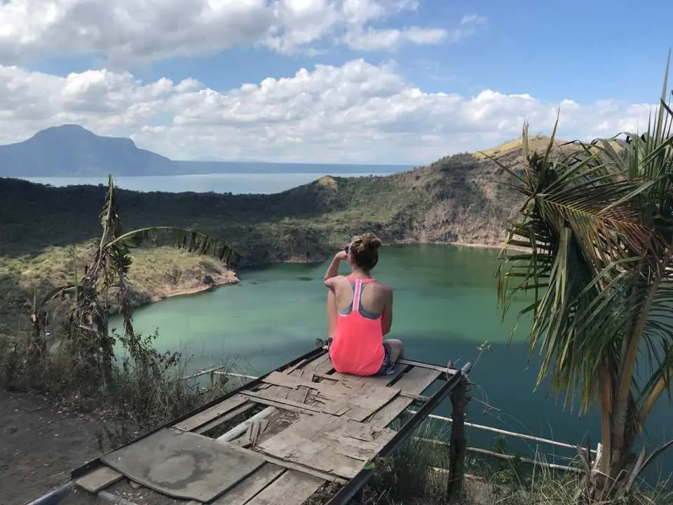 A visitor sitting on a viewing platform, overlooking Taal Volcano's emerald crater lake in the Philippines, with Taal Lake and rolling hills in the distance, offering expansive views.