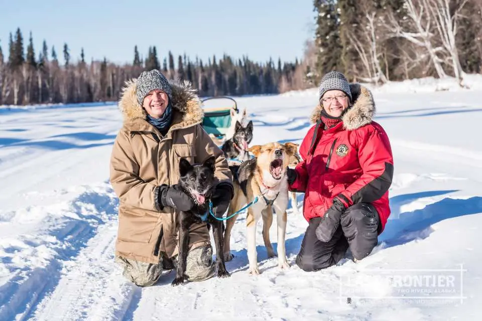 Two happy visitors kneeling in the snow, warmly interacting and petting friendly Alaskan huskies during a dog sledding tour in Fairbanks, Alaska, on a sunny winter day.