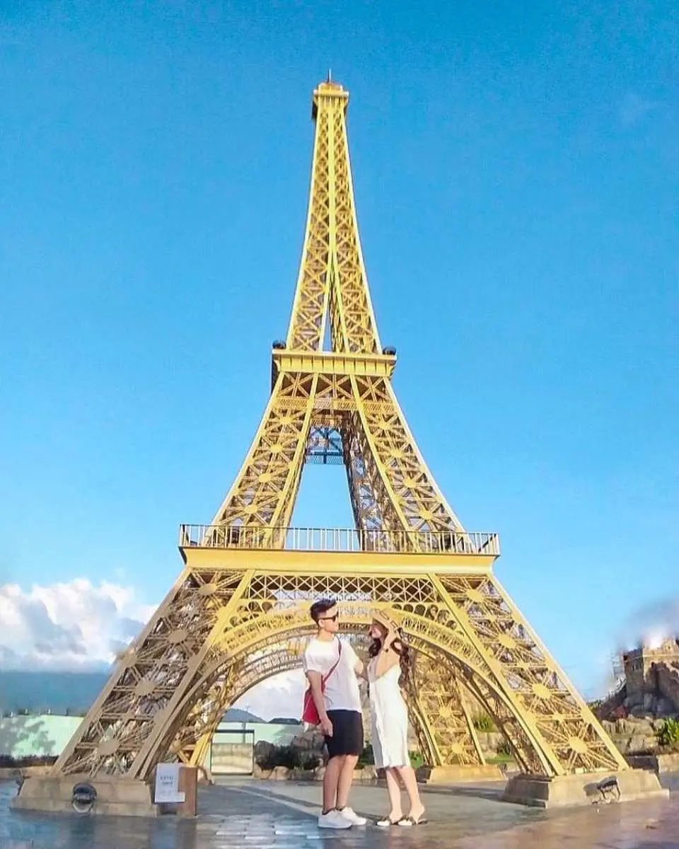 A couple poses romantically in front of a large golden replica of the Eiffel Tower in Da Nang under a clear blue sky, perfect for a memorable Instagram travel photo.