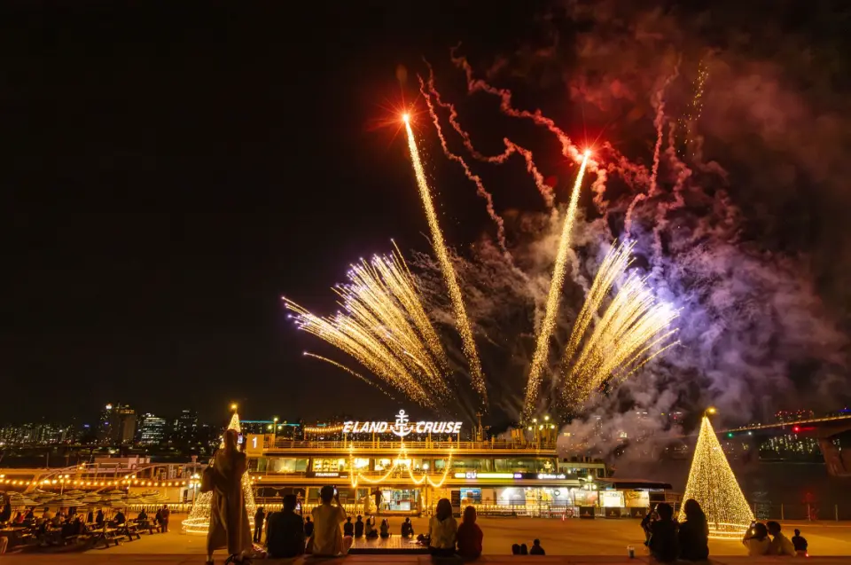 汝矣島衣戀遊輪碼頭，璀璨煙火點亮夜空，人們圍繞著發光的聖誕樹欣賞這場視覺盛宴。