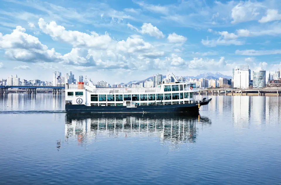 晴朗白天，衣戀遊輪在平靜漢江上航行，遊客盡覽首爾的城市風貌與藍天白雲。