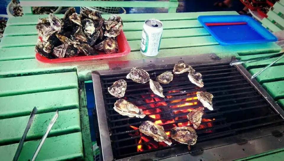 澎湖海洋牧場的戶外燒烤區，一整盤新鮮牡蠣和一瓶飲料放在綠色桌面上，烤爐上的牡蠣正滋滋作響，等待美味上桌。