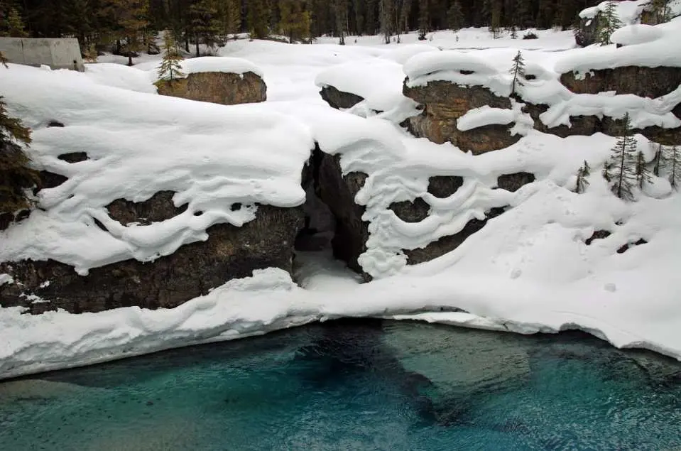 Winter scene of the Natural Bridge in Yoho National Park, where clear turquoise water flows beneath unique snow-covered rock formations, showcasing a distinct geological wonder.