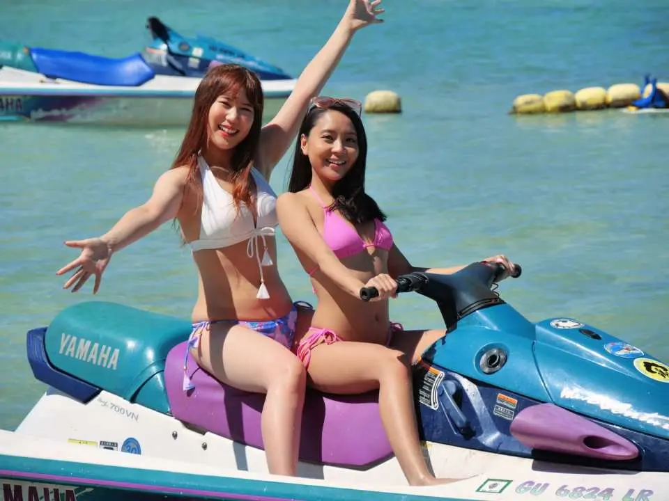 兩位年輕女性在關島清澈海域開心騎乘Yamaha水上摩托艇，揮手微笑，享受水上極速狂飆的樂趣。