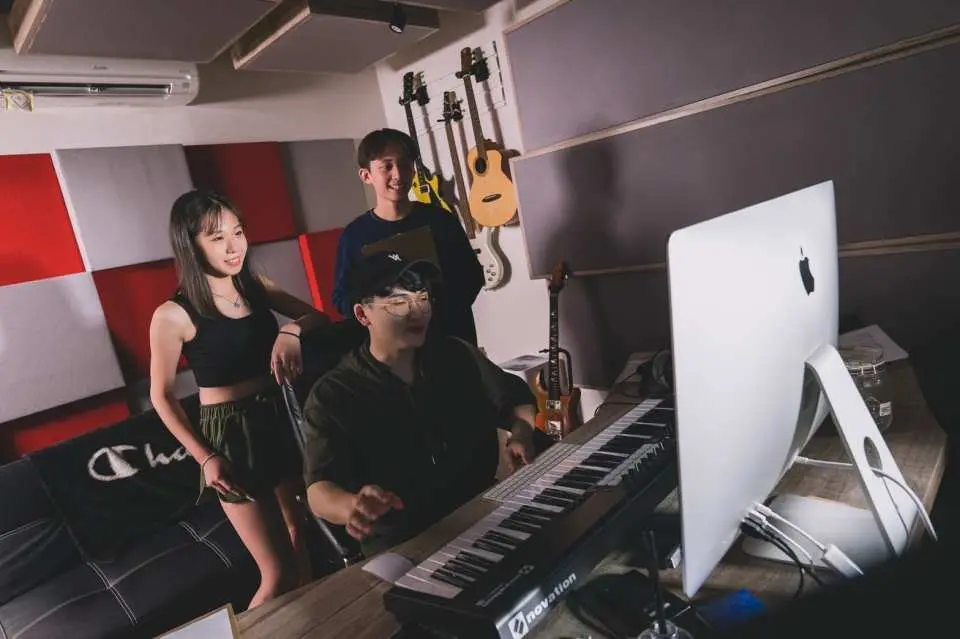 Two participants in a Taipei recording studio attentively watch an industry producer operate professional music production equipment and a keyboard, learning about the behind-the-scenes process of songwriting and recording.