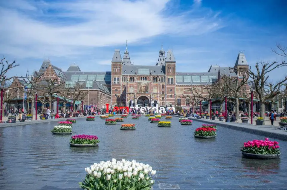 The iconic Rijksmuseum in Amsterdam, with 'I Amsterdam' sign and colorful tulips in reflecting pools.