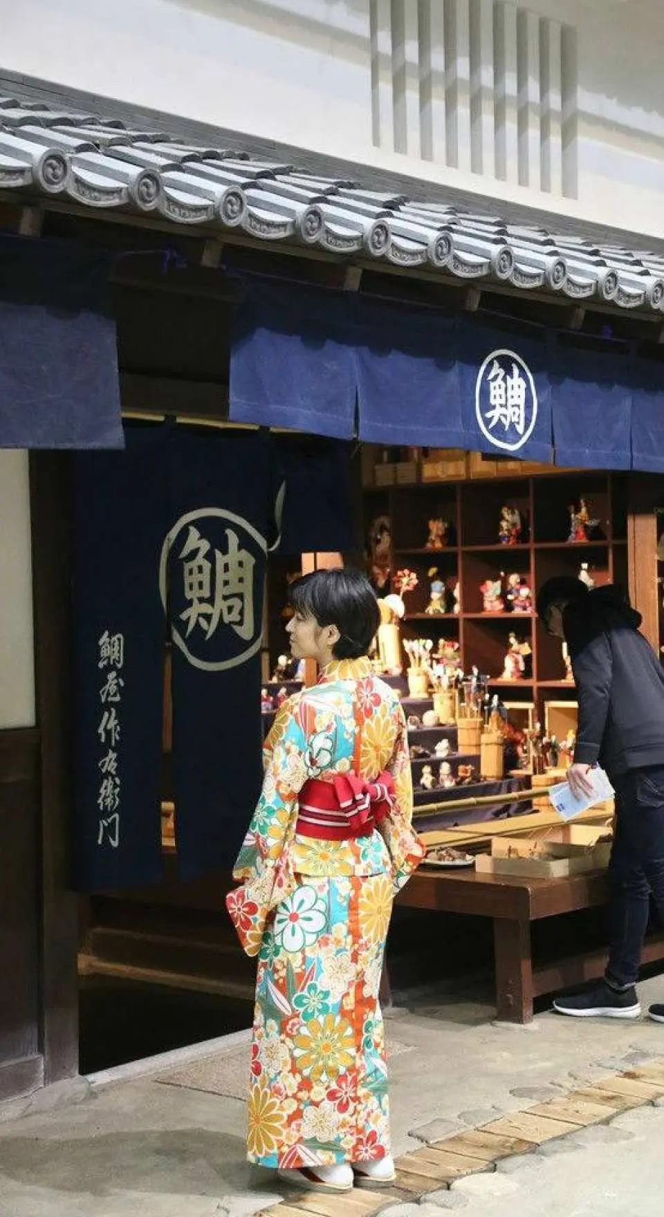 大阪今昔生活館內，遊客身著豔麗和服驕傲地站在江戶時代傳統商店前，參與沉浸式文化變裝體驗，讓歷史活現眼前。
