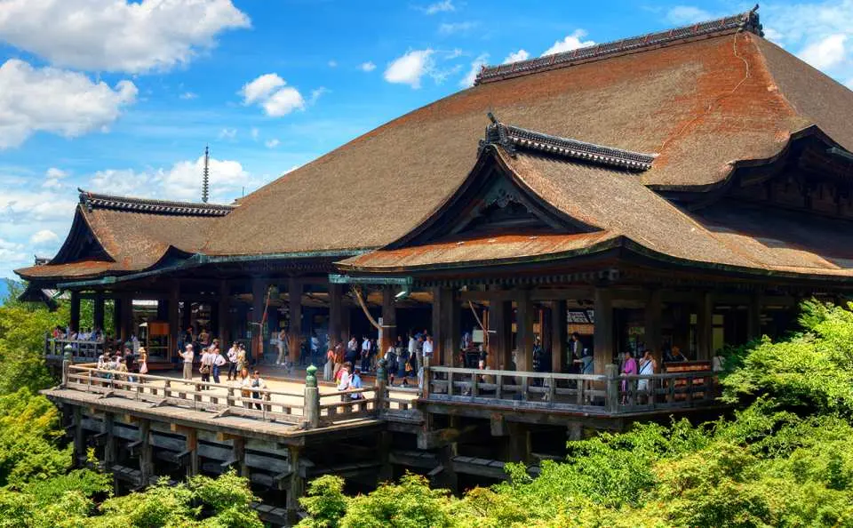Kiyomizu-dera Temple's famous wooden main hall in Kyoto, showcasing its unique cantilevered stage from above, surrounded by lush greenery, with visitors enjoying the scenic views and ancient temple atmosphere.