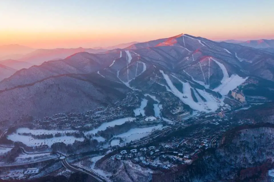 龙平度假村日出/日落航拍全景，雪白山峰染霞光，展现江原道冬季滑雪胜地令人屏息的梦幻景致。