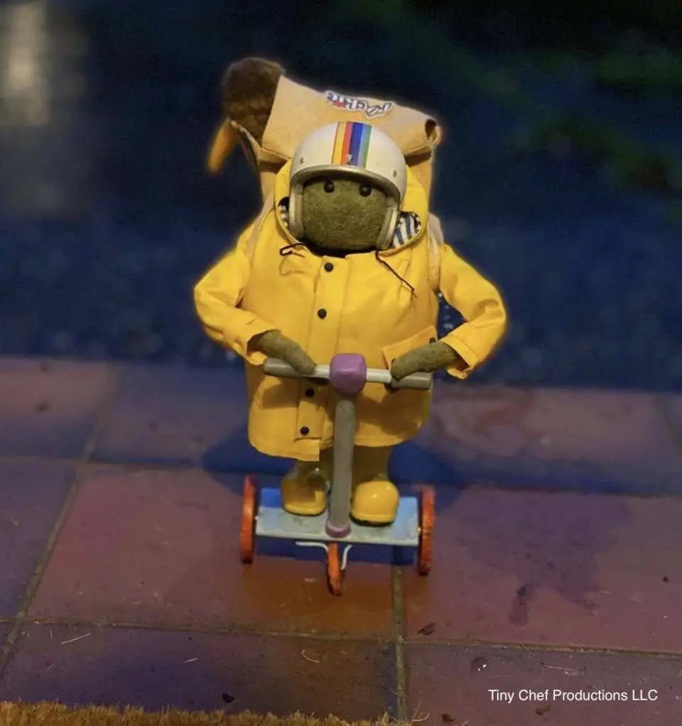 The distinctive frog puppet from California Academy of Sciences, wearing a yellow raincoat and helmet, rides a miniature scooter, showcasing its lively and playful character.