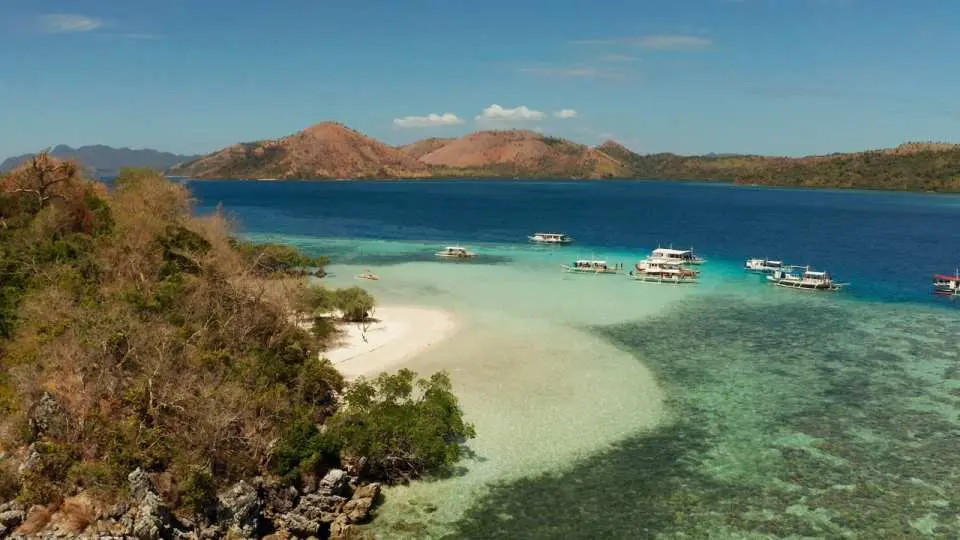 Serene tropical bay in Coron, featuring a wide expanse of shallow, crystal-clear turquoise waters and a small sandy area, surrounded by verdant hills and dry shrubs, with traditional boats gently bobbing, inviting visitors to experience island charm.